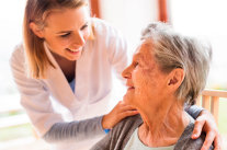 Pflegerin berührt Seniorin zum Gespräch an den Schultern. 